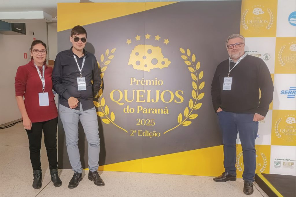 Estudante, usando óculos escuros, posando ao lado de duas pessoas — incluindo seu orientador — diante do painel do “Prêmio Queijos do Paraná 2025”, durante o evento.